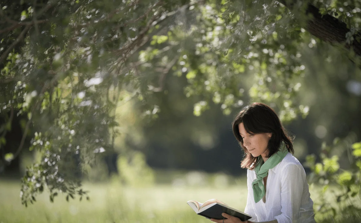 A woman enjoying leisure time reading under a tree, representing a calm lifestyle that can contribute to healthy aging skin.