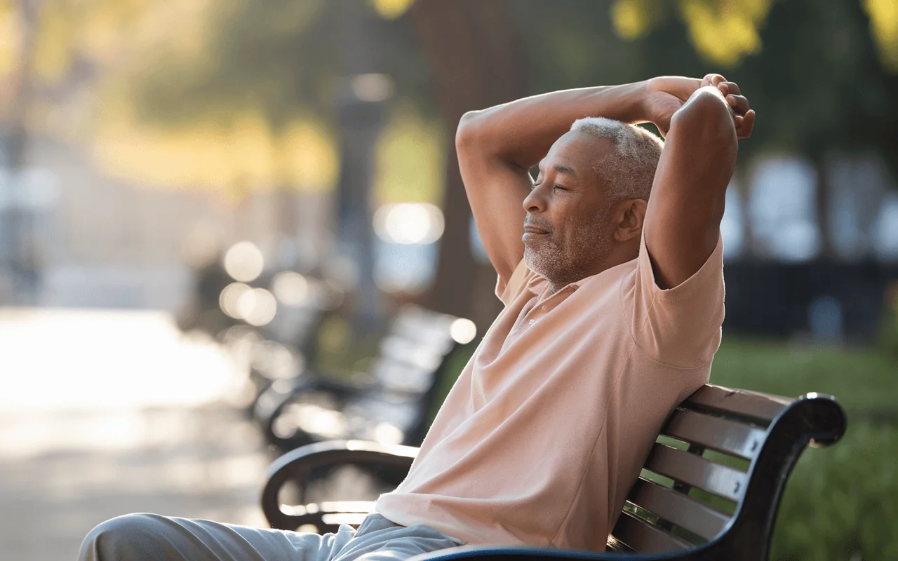 Older Black man with hands behind his head, eyes closed, relaxing on a park bench. Hope for arthritis pain relief.